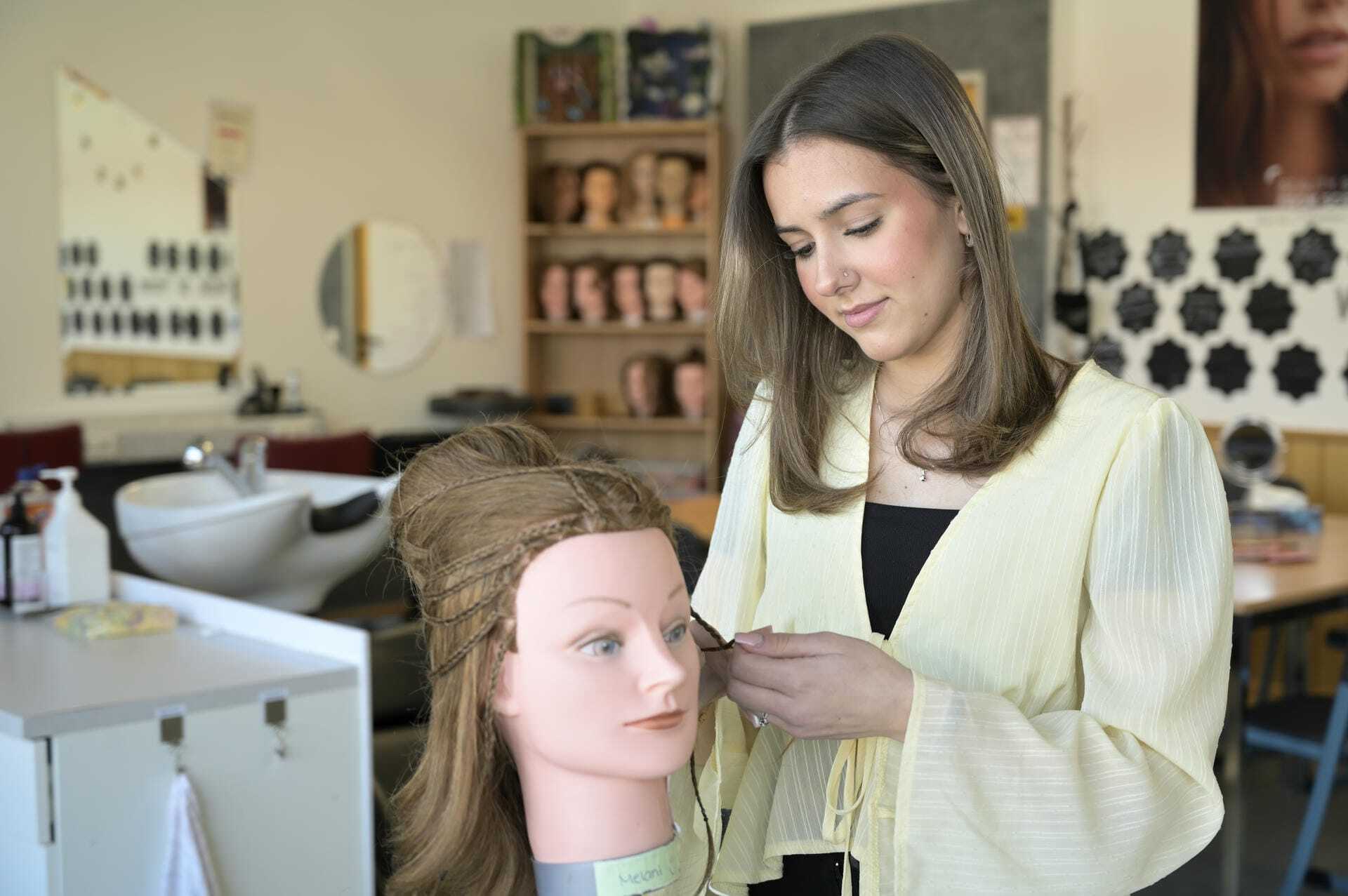 Eine Person arbeitet an einer Haarstyling-Puppe. Im Hintergrund sind weitere Puppen und Friseurequipment sichtbar.