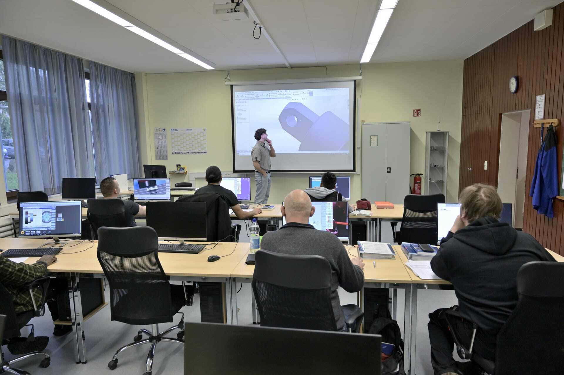 Ein Schulungsraum mit mehreren Teilnehmern an Computern. Ein Dozent präsentiert an einer Leinwand.